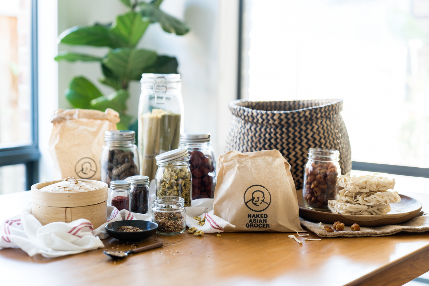 Table with dried ingredients, herbs, tea, noodles and paper bag with Naked Asian Grocer logo stamped on it